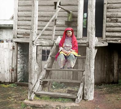 Eine baschkirische Frau sitzt in traditioneller Kleidung auf der Treppe vor einem Haus.