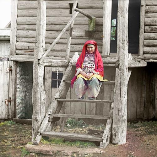 Eine baschkirische Frau sitzt in traditioneller Kleidung auf der Treppe vor einem Haus.