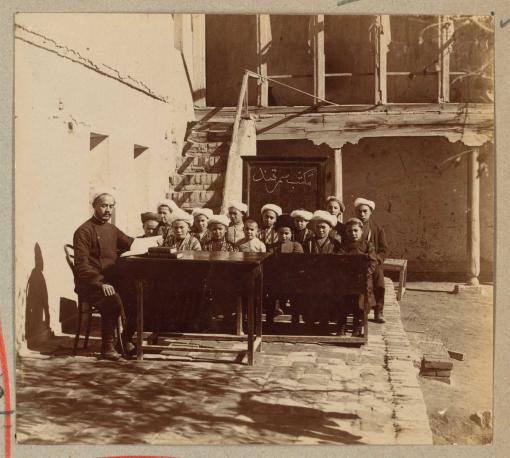 Ein usbekischer Lehrer und zwei Reihen junger männlicher Schüler an Tischen in einem Hof sitzend