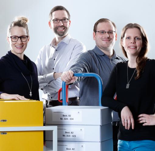 Teambild der Uni-Archiv Mitarbeiterinnen und Mitarbeiter mit Aktenkartons auf Lastkarren